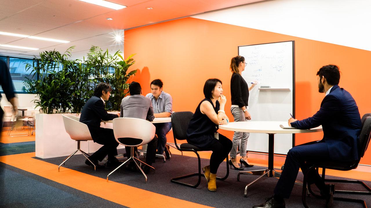 A woman writes on a white board, and people sit in groups around tables in a casual collaborative workspace with orange walls