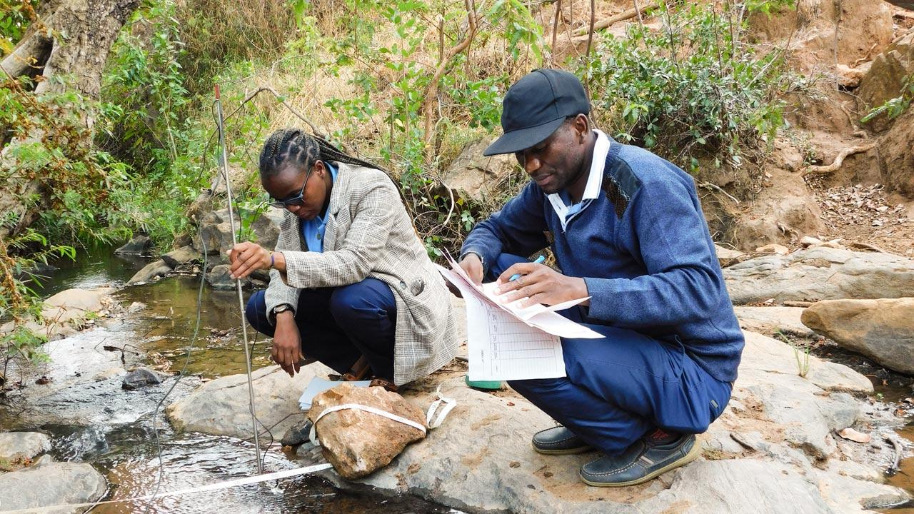 Two people sit on a rock beside a stream and measure the quality and quantity of the water